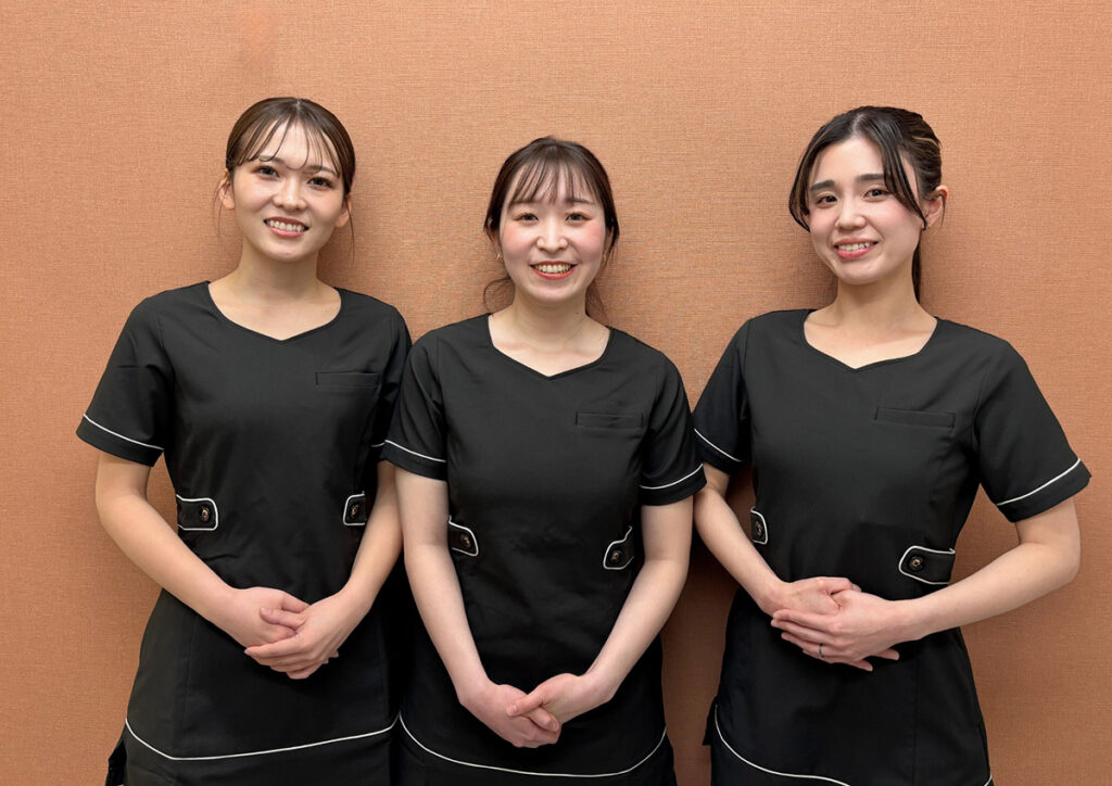 黒いユニフォームを着た3名のスタッフが、手を前に重ねて微笑みながら並んでいる